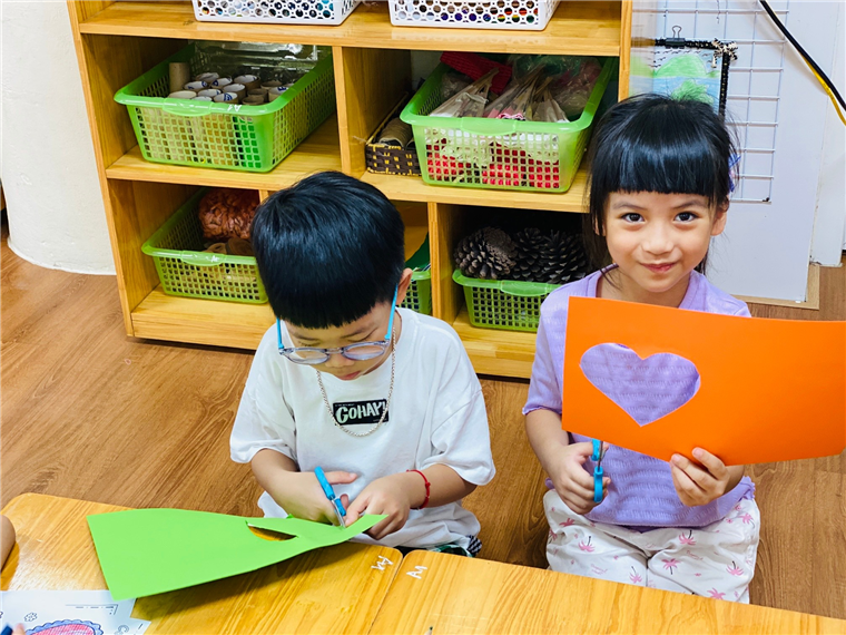 A couple of children sitting at a table with paper cutouts
Description automatically generated