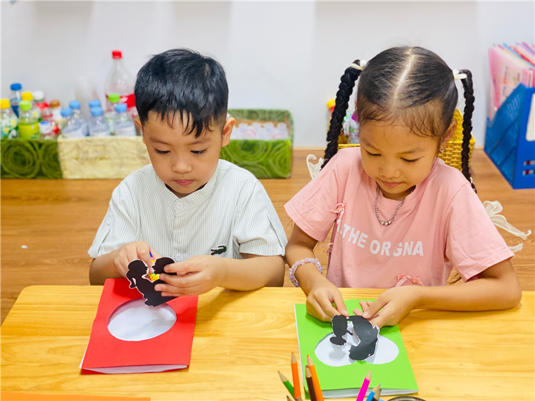 A child and child sitting at a table with paper cutouts
Description automatically generated