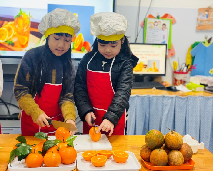 Two girls in aprons cutting orangesDescription automatically generated